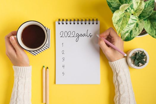 Desktop blank notepad and woman hands writing 2022 plans and goals text . Flat lay of yellow working table background with cup of tea and plants. Top view mock up