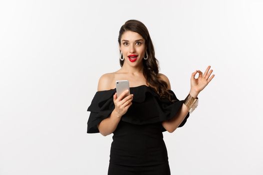 Online shopping concept. Attractive woman in trendy black dress, makeup, showing okay sign in approval and using mobile phone app, white background.