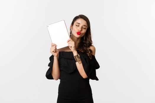 Fashion and shopping concept. Beautiful woman with red lips, wearing black dress, showing digital tablet and making kissing face, white background.