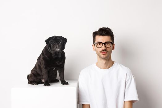 Image of handsome hipster in glasses sitting next to black cute pug dog, both staring at camera over white background.