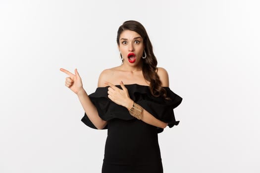 Fashion and beauty. Surprised woman in black glamour dress pointing fingers left, showing advertisement and staring amazed, white background.