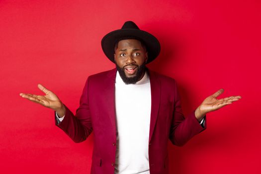 Image of confused Black man asking question, spread hands sideways and staring camera clueless, standing against red background.