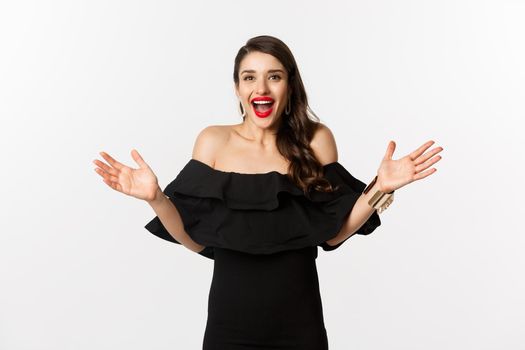 Beauty and fashion concept. Image of surprised and happy young woman in party dress reacting to good news, raising hands up and smiling amazed, white background.