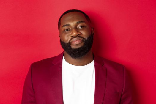 Close-up of handsome bearded plump guy smiling at camera shy, standing in stylish blazer on t-shirt, standing over red background.