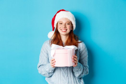 Winter holidays and Christmas Eve concept. Smiling redhead girl in sweater and Santa hat, holding New Year gift and looking at camera, standing against blue background.