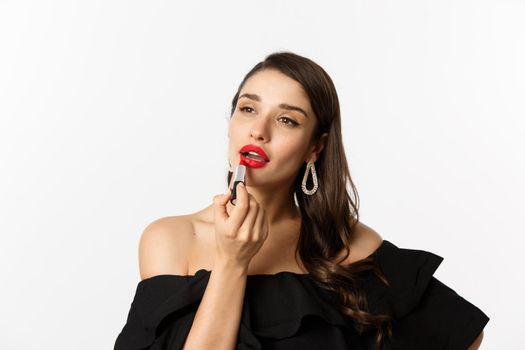 Fashion and beauty concept. Beautiful woman in black dress applying red lipstick and makeup, going on party, standing over white background.