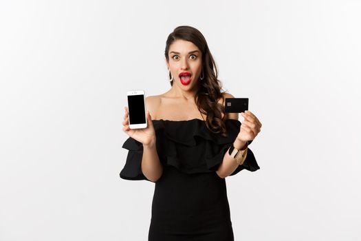 Fashion and shopping concept. Amazed beautiful woman showing smartphone screen and credit card, looking excited, buying online, white background.