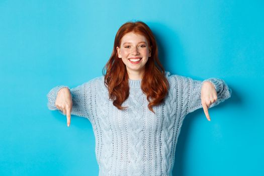 Winter holidays and people concept. Cheerful redhead woman in sweater pointing fingers down, smiling pleased at camera, showing promo, blue background.