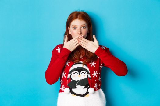 Winter holidays and Christmas Eve concept. Surprised redhead girl in cute sweater, gasping and covering mouth with hands, staring at camera, standing over blue background.