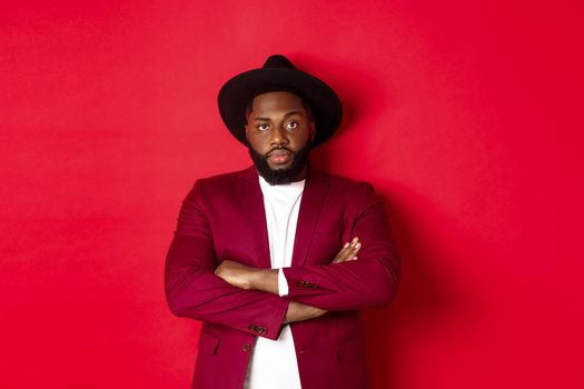 Serious black man standing in party outfit and waiting, cross arms on chest staring at camera, red background.