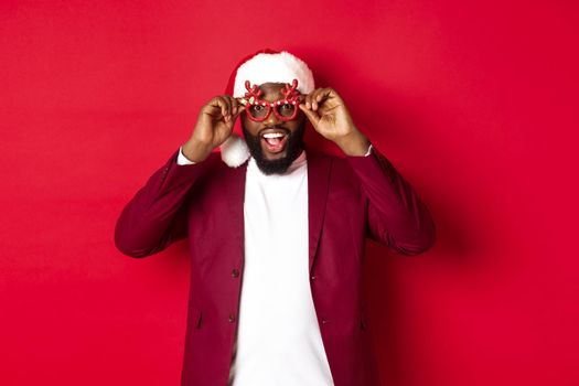 Funny Black man celebrating New Year, wearing party glasses and santa hat, having fun over red background.