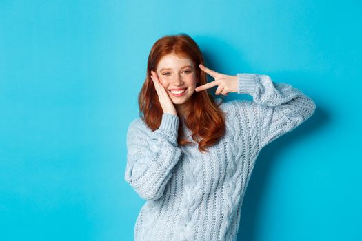 Cheerful redhead female model sending good vibes, smiling and showing peace sign, standing over blue background.