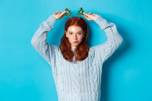 Winter holidays and Christmas sales concept. Beautiful redhead female model celebrating New Year, wearing funny party headband and sweater, smiling at camera.