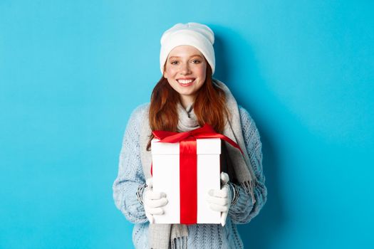 Winter holidays and Christmas sales concept. Cute redhead teeanage girl in beanie, sewater and scarf holding gift in wrapped box, smiling, wishing merry xmas, standing over blue background.