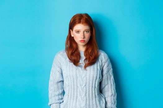 Sad and gloomy redhead teenage girl staring at camera uneasy, feeling bad, standing against blue backgorund in sweater.
