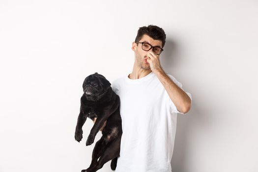 Young man shut nose as holding pug, disgusted with bad smell fart of animal, standing over white background.