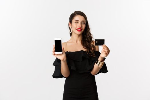 Fashion and shopping concept. Beautiful woman with red lips, winking at camera, showing smartphone screen and credit card, buying online, white background.