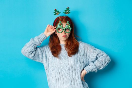 Winter holidays and Christmas sales concept. Beautiful redhead female model celebrating New Year, wearing funny party headband and glasses, smiling silly, blue background.