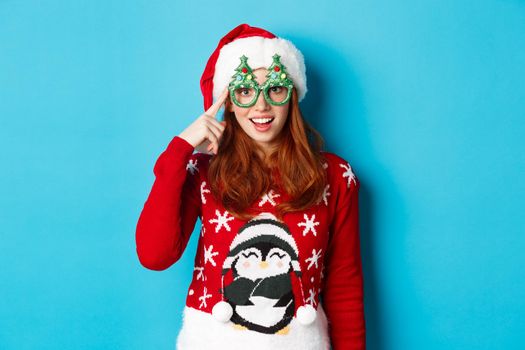 Happy holidays and Christmas concept. Funny redhead teen girl celebrating New Year, wearing santa hat and party glasses, standing against blue background.