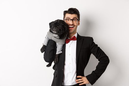 Handsome young man in suit and glasses holding cute black pug dog on shoulder, smiling happy at camera, wearing party outfits, white background.