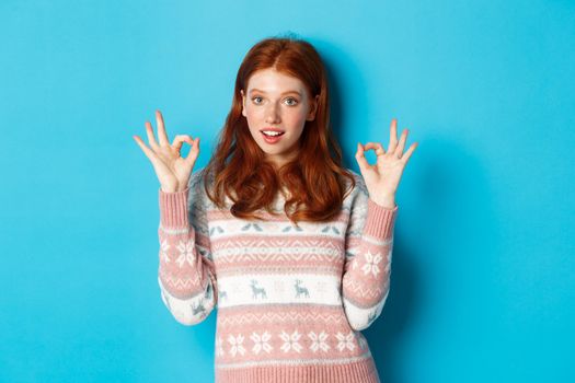 Image of young confident redhead woman showing okay signs, assure all good, praising choice, standing over blue background.