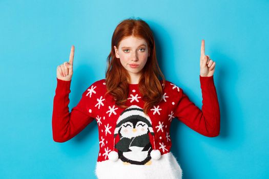 Merry Christmas. Serious and confident redhead girl pointing fingers up, showing advertisement and staring at camera.