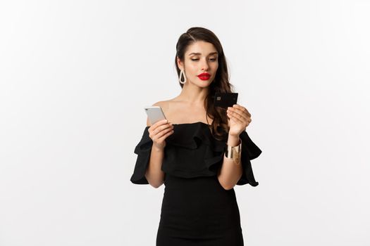 Fashion and shopping concept. Attractive woman in makeup and black dress order online, paying with credit card and mobile phone, white background.