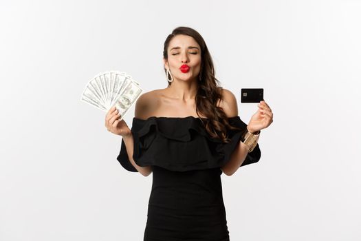 Beauty and shopping concept. Pretty glamour woman pucker lips for kiss, showing credit card and dollars, standing over white background.