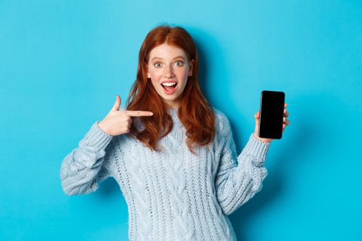 Impressed redhead girl pointing at phone screen, showing smartphone app or online offer and smiling excited, standing in sweater against blue background.