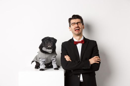 Animals, party and celebration concept. Sad dog owner crying, wearing suit, standing near cute black pug in costume, standing over white background.