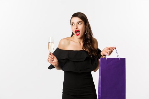 Excited woman shop and drink champagne, holding shopping bag, looking amazed, standing against white background.