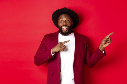 Winter holidays and shopping concept. Stylish african american man pointing fingers right at copy space for logo, standing over red background.