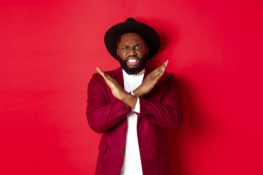 Angry Black man showing cross to stop something, looking furious and declining offer, standing in party clothes, red background.