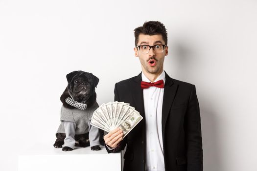 Amazed young man in party suit, standing near cute black pug dog in costume, entertained holding money dollars, standing over white background.