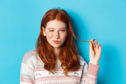 Close-up of cheeky redhead girl playing with hair strand, winking and smiling at camera, standing over blue background.