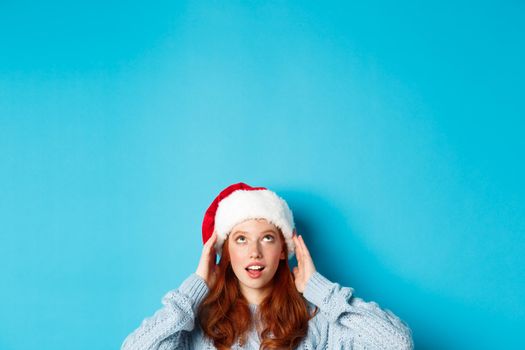 Winter holidays and Christmas eve concept. Head of cute redhead girl in santa hat, appear from bottom and looking up at copy space, staring logo, standing over blue background.