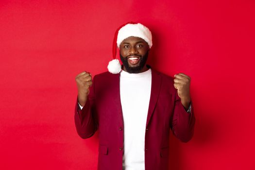 Christmas, party and holidays concept. Excited Black man celebrating New Year, wearing santa hat, clench fists satisfied, winning and rejoicing, standing against red background.