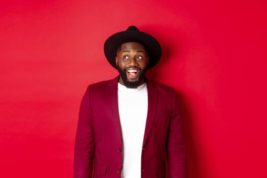 Fashion and party concept. Excited african american man looking at logo, staring upper left corner with happy smile, standing against red background.