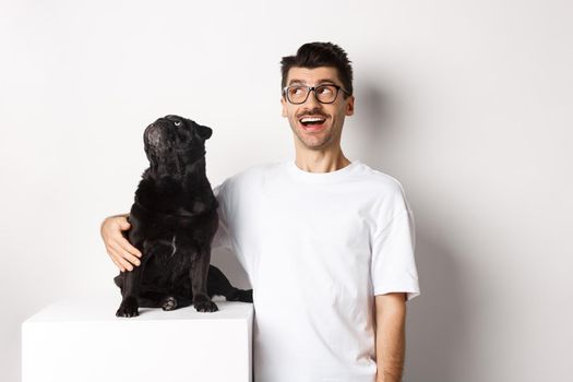 Amazed young man in glasses hugging his dog, pet owner and pug staring at upper left corner promo offer, standing over white background.