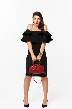 Beauty and fashion concept. Full length if silly young woman pouting and looking surprised, holding purse, wearing heels and black dress, white background.