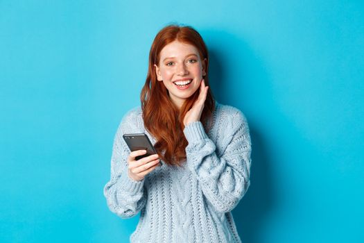 Beautiful redhead girl in sweater, smiling at camera, using mobile phone app, standing with smartphone against blue background.