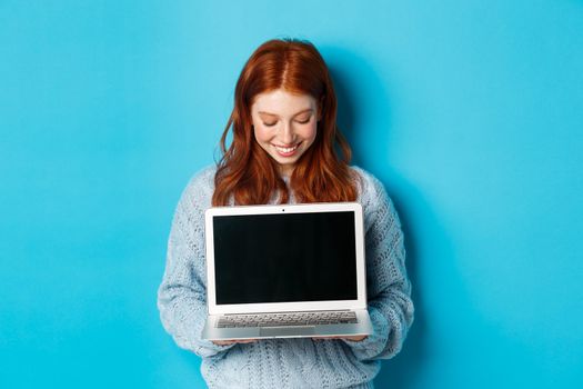 Cute redhead woman in sweater, showing and looking at laptop screen with pleased smile, demonstrating something online, standing over blue background.