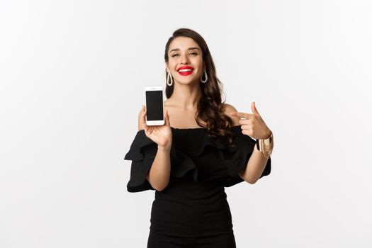 Online shopping concept. Fashionable woman in black dress pointing finger at smartphone screen, showing application, standing over white background.