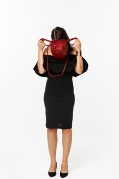 Beauty and fashion concept. Full length of young woman sticking head inside purse and searching something, wearing black dress and high heels, standing over white background.