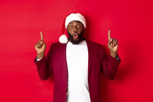 Christmas, party and holidays concept. Impressed Black man showing offer, pointing fingers up at logo, saying wow, standing against red background.