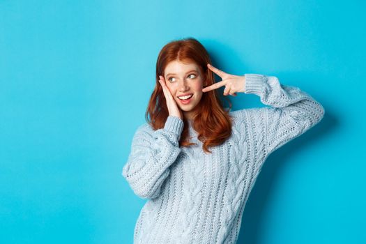 Happy redhead girl smiling, showing peace sign and looking left at promo, standing in sweater against blue background.