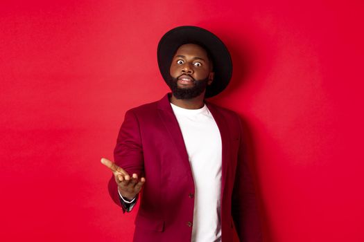 Shocked african american man staring at camera and pointing at something upsetting, standing in party jacket and hat, red background.