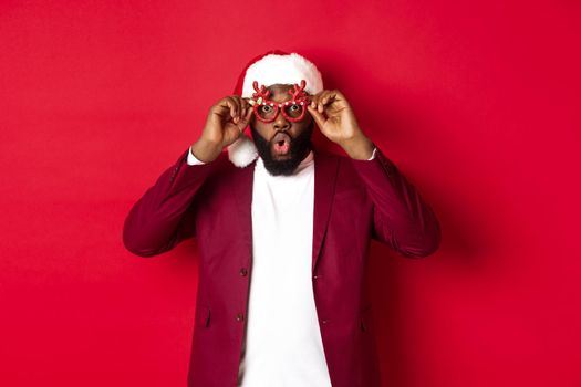 Funny Black man celebrating New Year, wearing party glasses and santa hat, having fun over red background.