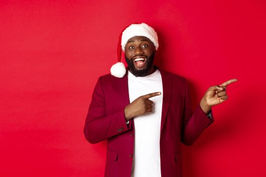Christmas, party and holidays concept. Cheerful Black man smiling, pointing fingers right and showing advertisement, standing over red background.