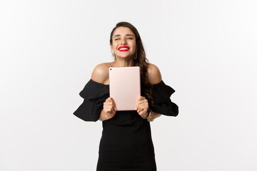 Fashion and shopping concept. Happy young woman with red lips, wearing black dress, rejoicing and holding digital tablet, winning prize, white background.
